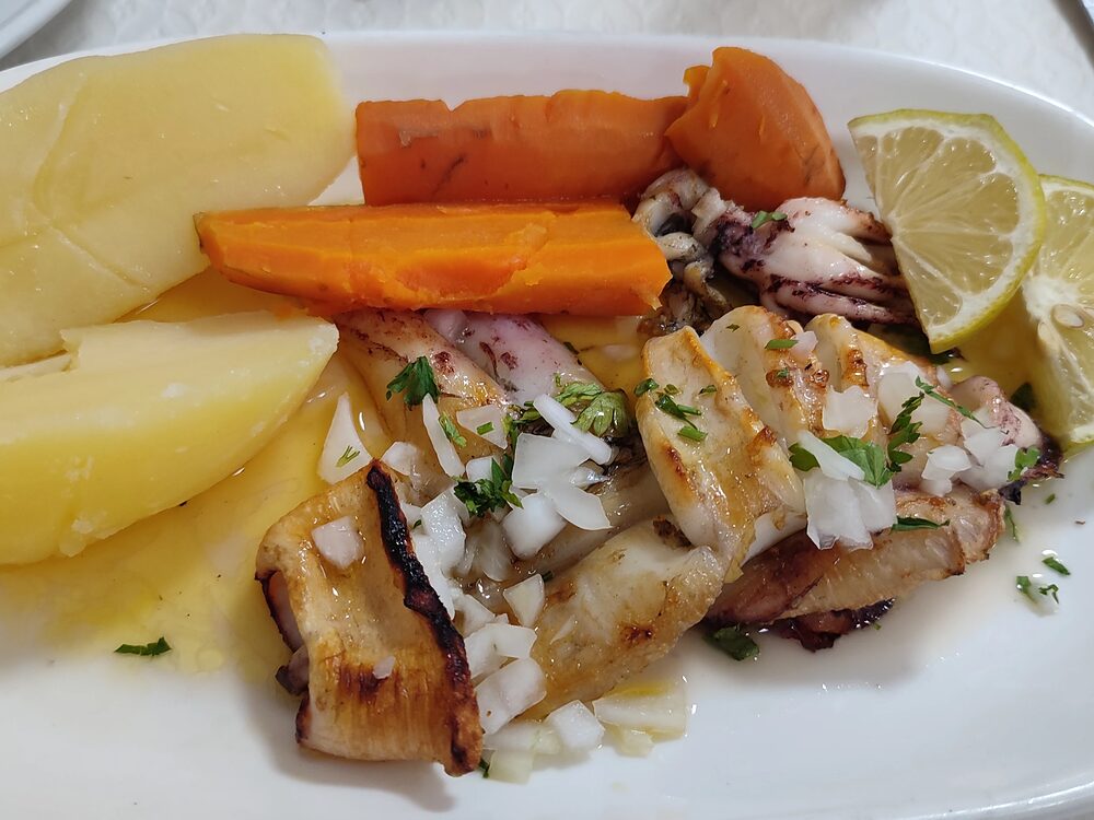 Choco grelhado às tiras num prato branco, com batata cozida, cenoura, cebola picada e limão