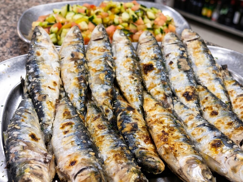 Travessa de metal cheia de sardinhas assadas, com uma salada de tomate e pepino atrás