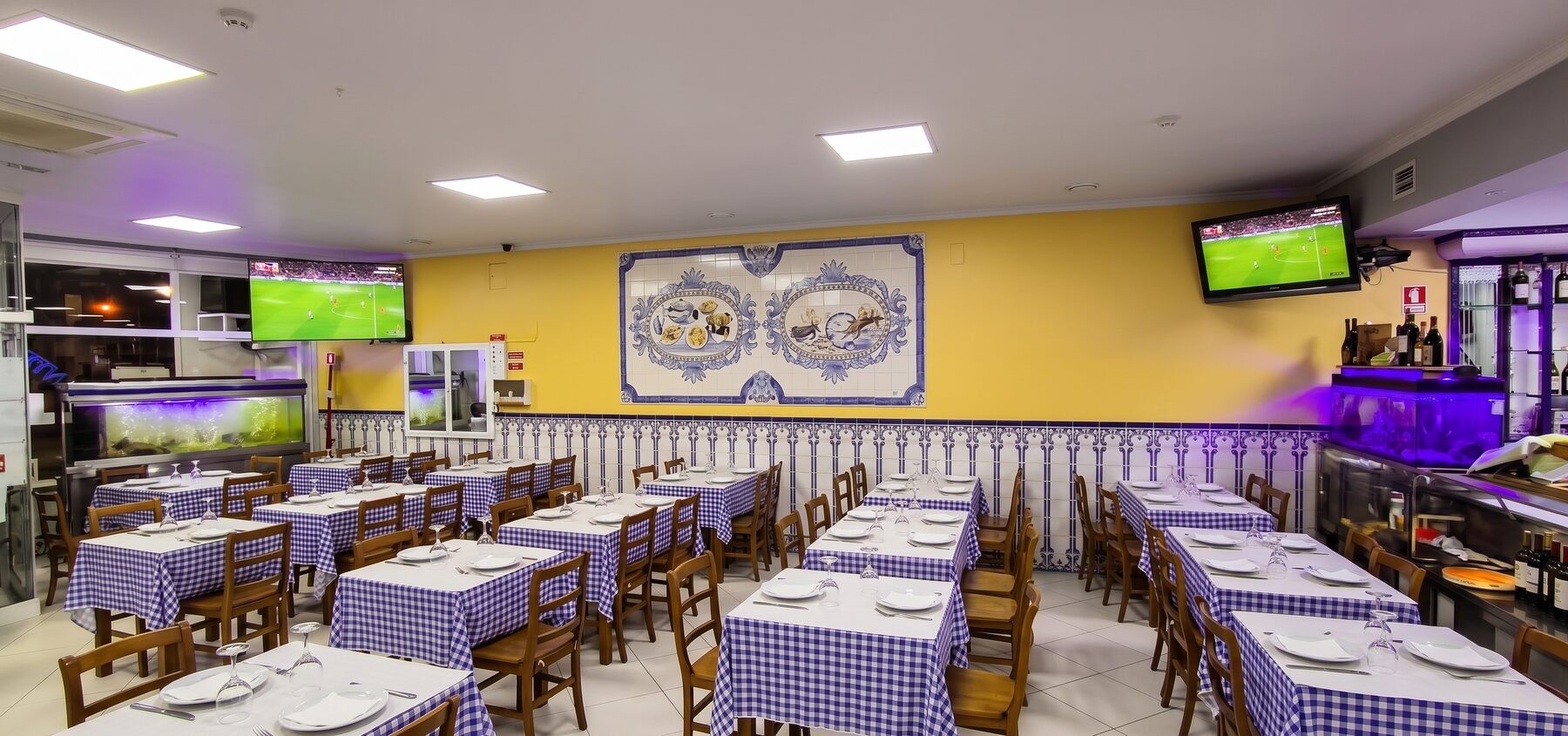 Sala da marisqueira com mesas de toalha de xadrez azul, painel de azulejo azul e branco na parede amarela e viveiros de marisco ao fundo